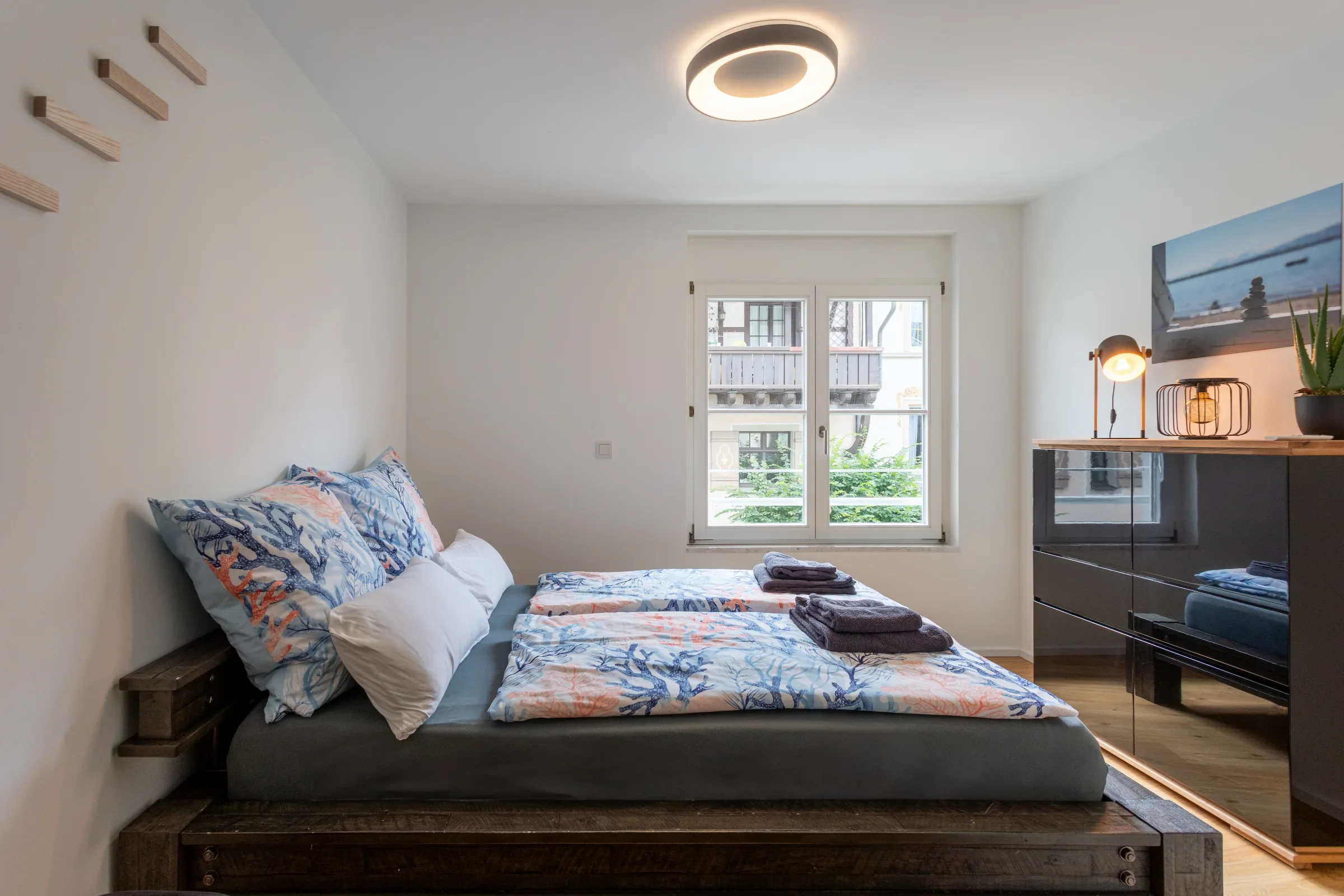 Kleines Schlafzimmer mit Doppelbett, gemusterten Bettbezügen, Fenster und Kommode mit Lampe